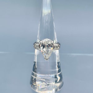 May include: A silver ring with a large, pear-shaped clear stone as the centerpiece. Two smaller, rectangular stones flank the main stone. The ring is displayed on a clear, conical stand against a light blue background.