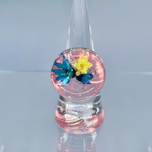 May include: A pink resin ring with a clear dome containing blue and yellow pressed flowers and glitter.