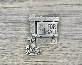 May include: A silver-toned metal charm depicting a house for sale sign with flowers. The sign reads "FOR SALE" and "SOLD".