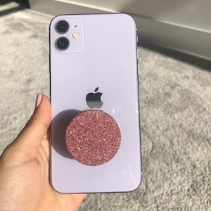 May include: A white iPhone with a pink glitter phone grip attached to the back.