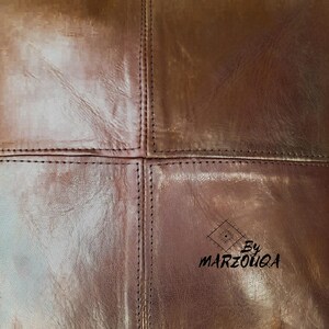 May include: Close-up of brown leather with visible stitching. The leather is divided into four sections with detailed stitching along the seams. The text "By MARZOUQA" is visible in the lower right corner.