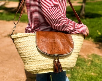 May include: Woven straw shoulder bag with a brown leather flap and tassel detail. The bag has a long brown strap and the text "ByMarzouga" is visible on the flap. The bag is carried by a person wearing a red and white striped shirt.