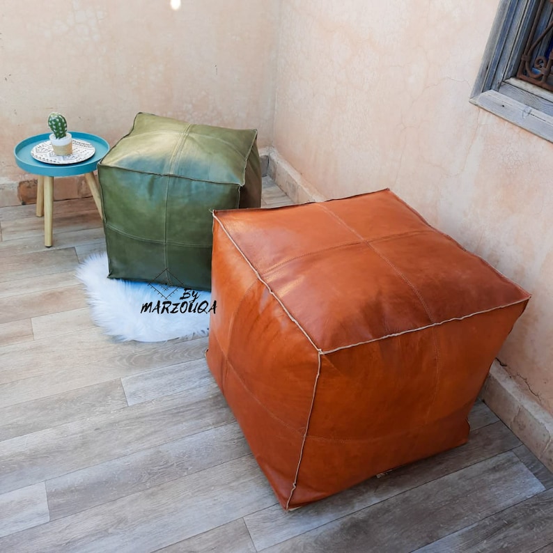 May include: Two leather poufs, one olive green and one rust-colored, sit on a wooden floor. The square poufs have stitched panels and white stitching. A small blue side table with a cactus is in the background.