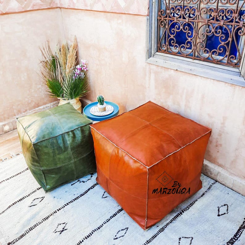 May include: Two square leather poufs, one olive green and one rust-colored, sit on a patterned rug. The rust-colored pouf has the text "BY MARZOUGA" on its side. A small blue table with a cactus and a vase of plants are in the background.