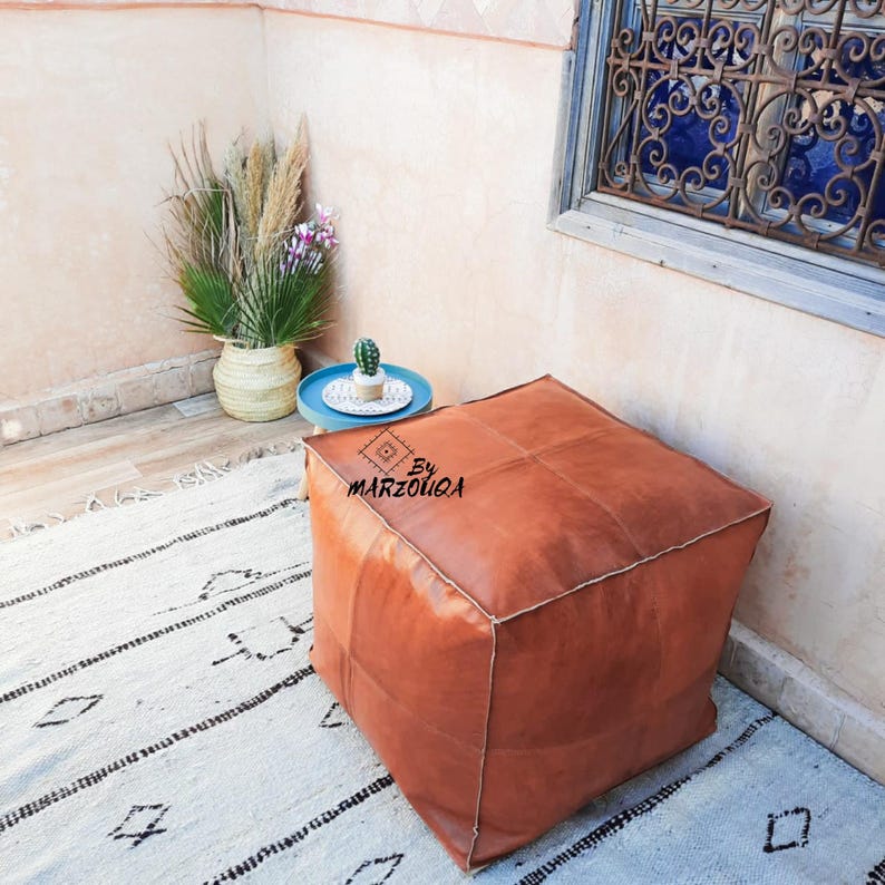 May include: A square, brown leather ottoman with stitched panels. The ottoman is placed on a white rug with black geometric patterns. A small blue table with a cactus and a vase with plants are in the background. The text "By MARZOUQA" is on the ottoman.