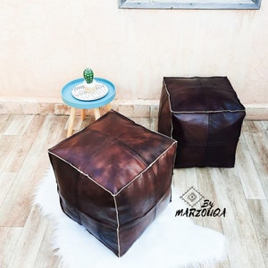 May include: Two dark brown leather cube ottomans with white stitching. The ottomans are placed on a white faux fur rug. A small blue side table with a cactus and a decorative mat is in the background. The text "By MARZOUQA" is in the bottom right corner.