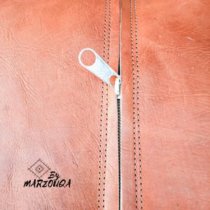 May include: Close-up of a brown leather surface with a silver zipper partially open. The leather has visible grain and is stitched with a contrasting thread. The zipper pull is a simple, functional design. The text "By MARZOUQA" is visible.