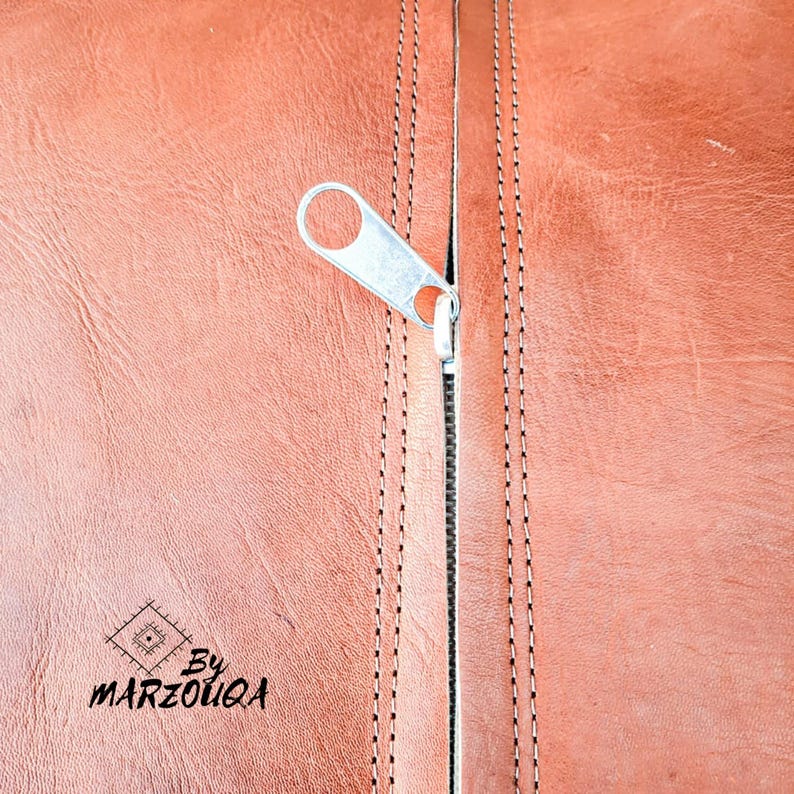 May include: Close-up of a brown leather surface with a silver zipper partially open. The leather has visible grain and is stitched with a contrasting thread. The zipper pull is a simple, functional design. The text "By MARZOUQA" is visible.