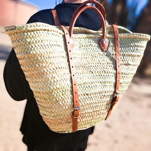 May include: A woven straw tote bag with brown leather straps and handles. The bag has a wide opening and a rectangular base. The leather straps are attached to the bag with metal buckles. The bag is carried over the shoulder.