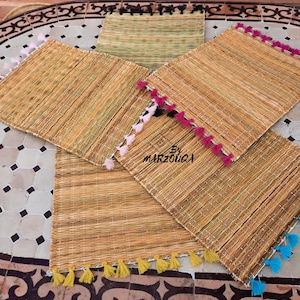 Op de afbeelding: Een verzameling geweven placemats met kleurrijke kwastjes. De placemats zijn rechthoekig en gemaakt van natuurlijke vezels, met kwastjes in roze, zwart, geel en blauw. De tekst "By MARZOUGA" is zichtbaar.