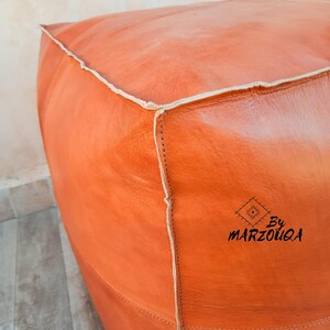 May include: A square, orange leather ottoman with white stitching along the edges. The ottoman has a textured surface and a logo that reads "By MARZOUQA" in black script.