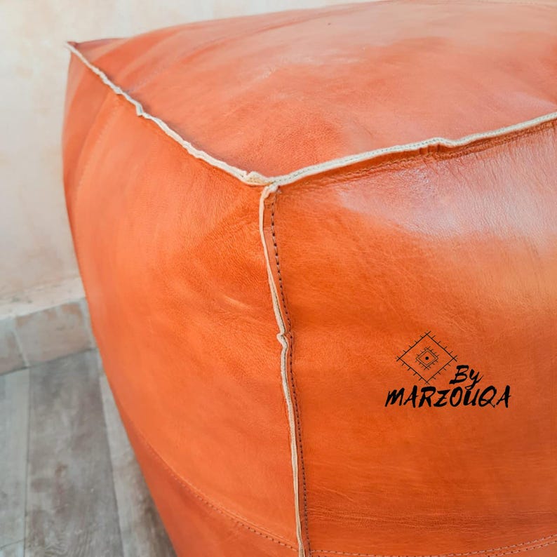 May include: A square, orange leather ottoman with white stitching along the edges. The ottoman has a textured surface and a logo that reads "By MARZOUQA" in black script.