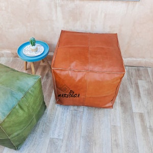 May include: A square, brown leather ottoman with white stitching and a stitched design. The ottoman is part of a set, with a green ottoman visible in the background. A small blue table with a cactus is also in the background. The text "By MARZOUQA" is visible.