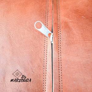 May include: Close-up of a brown leather surface with a silver zipper and decorative stitching. The zipper is partially open, revealing the interior. The text "By MARZOUQA" is visible in the lower left corner.