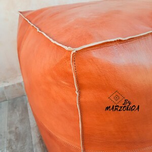 May include: Orange leather pouf with white stitching and the text "By Marzouqa". The square-shaped ottoman has a smooth, textured surface. The stitching is visible along the edges, and the leather has a slight sheen.