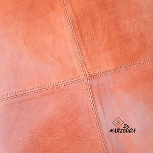 May include: Close-up of a section of a leather product in a warm, reddish-brown hue. The leather is stitched together in a geometric pattern with dark thread. The surface has a textured appearance, with visible grain and subtle variations in color.
