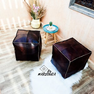 May include: Two dark brown leather cube ottomans with white stitching. One ottoman sits on a white faux fur rug. A small blue table with a cactus and a woven vase with dried plants are in the background. The text "By MARZOUQA" is on the rug.