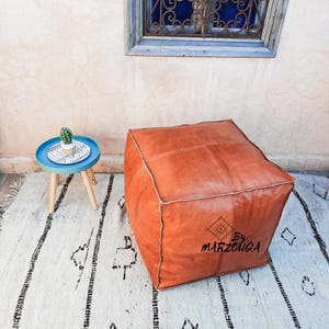 May include: A large, square, orange leather pouf with white stitching. The pouf sits on a white rug with black geometric patterns. A small blue side table with a cactus and a decorative tray is to the left.