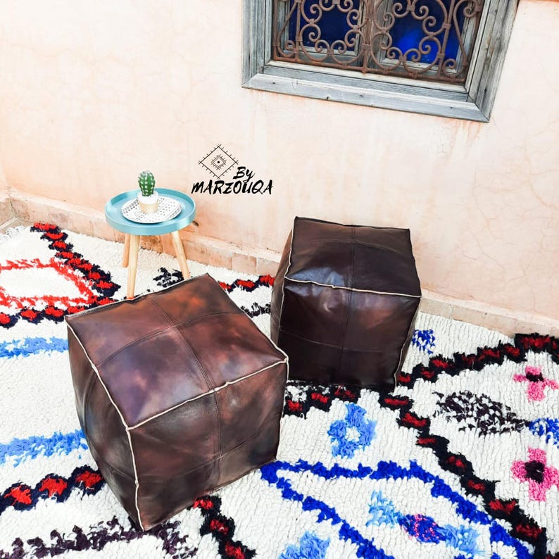 May include: Two brown leather ottomans with white stitching sit on a colorful rug with blue, red, and pink patterns. A small blue table with a cactus sits in the background. The image is taken outdoors.
