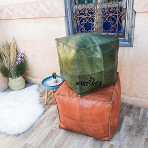 May include: Two square leather ottomans, one olive green and one rust-colored, stacked on a wooden floor. The green ottoman has the text "By MARZOUOA" on it. A small blue table with a cactus and a white rug are in the background.