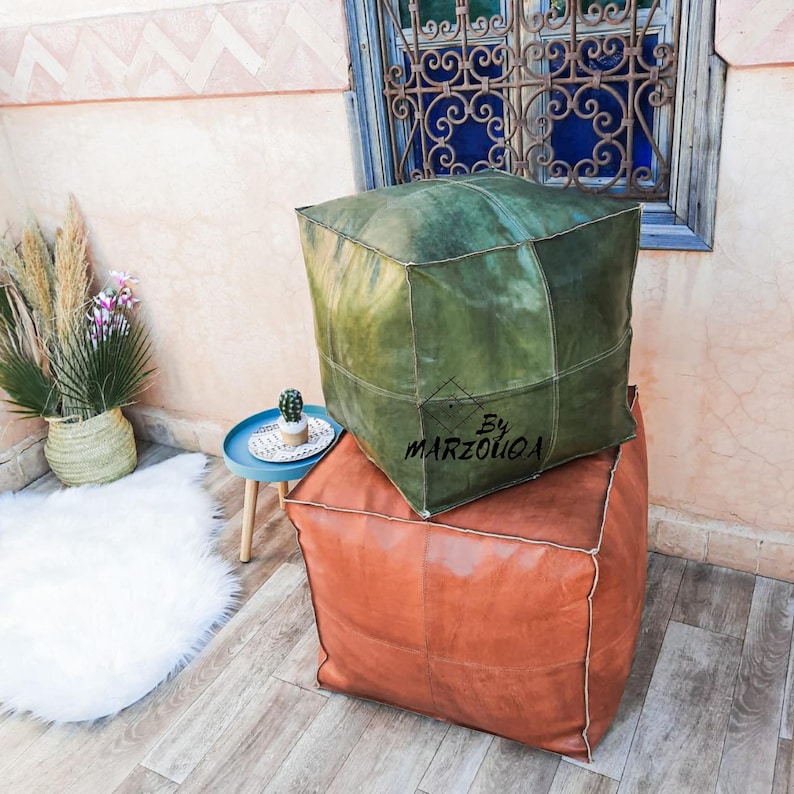 May include: Two square leather ottomans, one olive green and one rust-colored, stacked on a wooden floor. The green ottoman has the text "By MARZOUOA" on it. A small blue table with a cactus and a white rug are in the background.