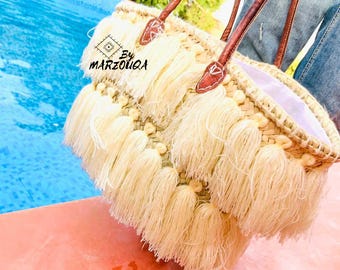 May include: Cream-colored straw tote bag with long, cream-colored fringe and brown leather handles. The bag has a woven design and the text "By MARZOUQA" is visible on the front. The bag is near a pool.