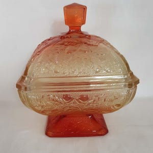 May include: A square orange glass candy dish with a matching lid featuring a square finial. The dish has a pedestal base and is decorated with embossed leaf and acorn patterns on the sides and lid, showcasing a gradient of deep orange to light amber.