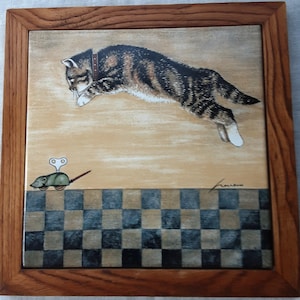 May include: A framed ceramic tile featuring a tabby cat leaping towards a wind-up mouse. The cat is brown and black with a brown collar. The background is tan with a checkered pattern at the bottom. The frame is wood.