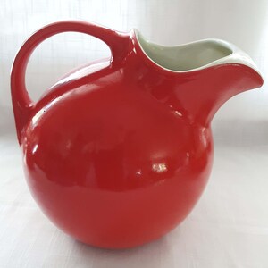 May include: A bright red ceramic pitcher with a spherical body, a curved handle on the left side, and a wide, smooth pouring spout. The interior of the pitcher is finished in a contrasting off-white glaze, visible through the open top opening.