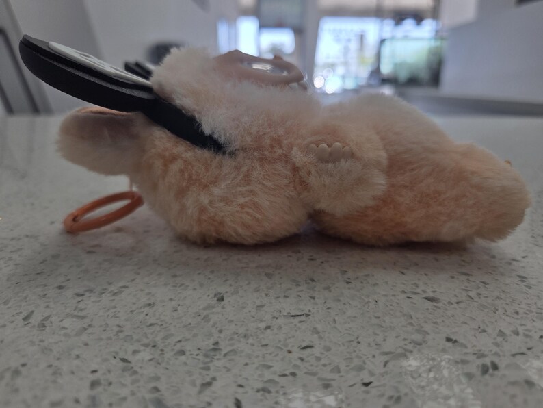 May include: A plush, peach-colored key holder shaped like a small animal, with a black key inserted. The key holder has a small orange ring attached. The plush material appears soft and fuzzy.