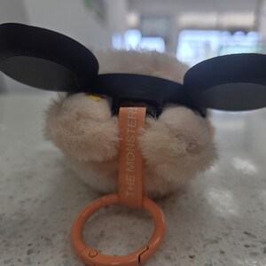 May include: A plush toy with large black ears and a light brown, furry body. The toy has a small orange tag that reads "THE MONSTER" and an orange clip. The toy is resting on a speckled surface.
