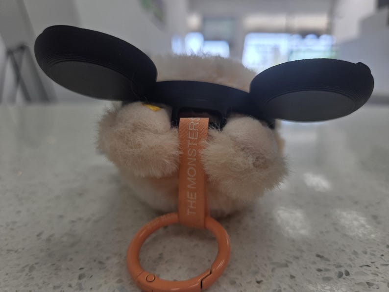 May include: A plush toy with large black ears and a light brown, furry body. The toy has a small orange tag that reads "THE MONSTER" and an orange clip. The toy is resting on a speckled surface.