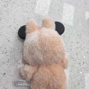 May include: A plush, peach-colored bear-shaped phone case with black circular ears. The case has a soft, furry texture and is designed to fit a smartphone. The bear's back and bottom are visible in the image.