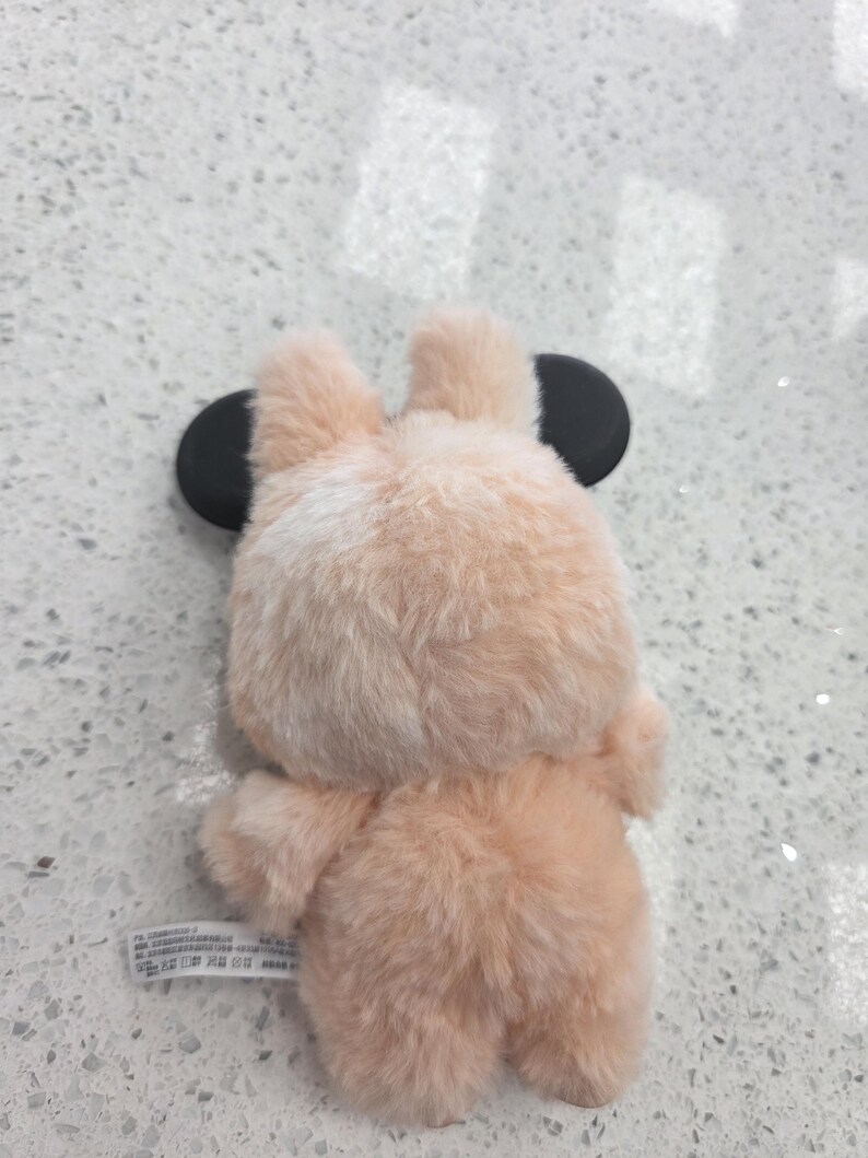 May include: A plush, peach-colored bear-shaped phone case with black circular ears. The case has a soft, furry texture and is designed to fit a smartphone. The bear's back and bottom are visible in the image.
