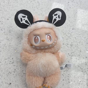 May include: A plush toy in a light peach color, featuring a smiling face and a furry body. The toy has large, round ears with a Scream mask design. The toy's eyes are oval-shaped and the mouth has visible teeth.