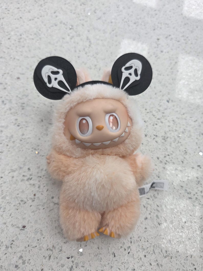 May include: A plush toy in a light peach color, featuring a smiling face and a furry body. The toy has large, round ears with a Scream mask design. The toy's eyes are oval-shaped and the mouth has visible teeth.