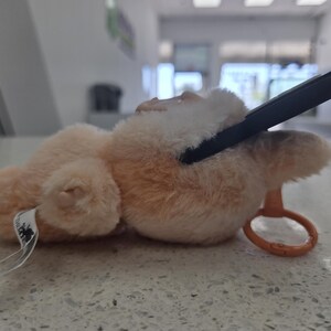 May include: A plush, light brown teddy bear with a black rectangular object resting on its head. The bear has a small orange ring attached. A white tag with black text is attached to the bear's body.