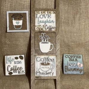 May include: A set of five wooden signs with coffee-themed sayings. The signs are painted in various shades of brown, blue, and white. The signs feature text such as "This home runs on love, laughter, and lots of strong coffee", "Life begins coffee helps", "Coffee makes me love murdery", and "Coffee bar love is brewing".