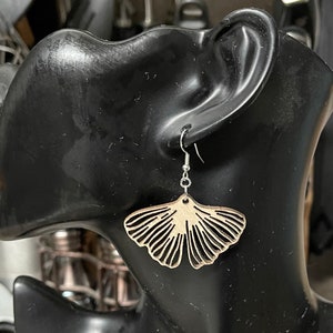May include: A pair of wooden earrings shaped like ginkgo leaves. The earrings are a light brown color and have a delicate, intricate design. They are hanging from silver hooks.