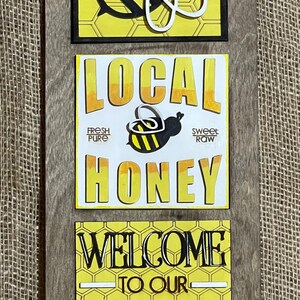 May include: A wooden sign with three panels. The top panel features a black and white bee with yellow honeycomb. The middle panel says "LOCAL HONEY" with a smaller bee image and the words "FRESH PURE" and "SWEET RAW". The bottom panel says "WELCOME TO OUR HIVE" with a yellow honeycomb background.