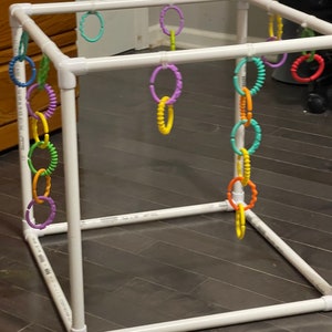 May include: A white plastic frame with colorful plastic rings hanging from it. The rings are in various colors, including yellow, orange, red, green, blue, and purple. The frame is made of PVC pipe and is designed to be used as a baby play gym.
