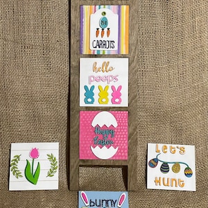 May include: Wooden ladder with five signs for Easter decorations. The signs have different colours and designs, including carrots, bunnies, eggs, and the words "hello peeps", "happy easter", "let's hunt", and "bunny trail".