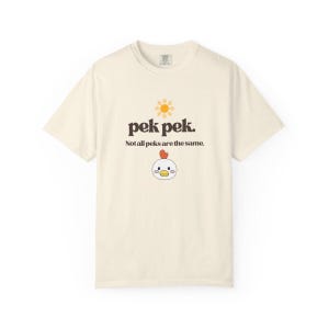 Könnte beinhalten: Cremefarbenes T-Shirt mit dem Text "pek pek." und dem Satz "Not all peks are the same." Eine Cartoon-Sonne und ein Hühnergesicht sind ebenfalls abgebildet. Das T-Shirt ist ein Freizeitartikel.