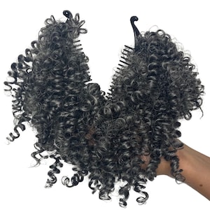 May include: A dark gray curly hairpiece with a black comb clip. The hairpiece is styled in voluminous, tight curls. The hairpiece is held against a white background.