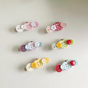 Pode incluir: Seis ganchos de cabelo com bases em forma de maçã decoradas com vários botões coloridos e pequenos detalhes em forma de flor ou coração. Os ganchos apresentam uma variedade de padrões de xadrez vichy em tons de rosa, roxo, verde, amarelo e azul.