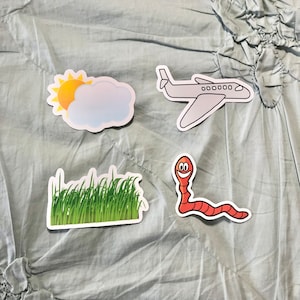 Puede incluir: Cuatro pegatinas coloridas sobre un fondo de tela azul claro. Las pegatinas son un sol y una nube, un avión, un parche de hierba verde y un gusano rojo con una cara sonriente.