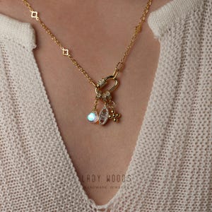 Puede incluir: Un collar de cadena dorada con un dije en forma de corazón y varios dijes colgantes, incluyendo un cristal transparente, una cruz y una pequeña perla dorada. El collar está sobre un fondo blanco con el texto "LADY WOODS HANDMADE JEWELRY".