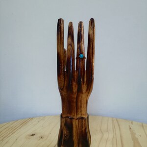 K&ouml;nnte beinhalten: Ein handf&ouml;rmiger Ringhalter aus Holz mit dunkelbraunem Finish. Ein t&uuml;rkisfarbener Ring wird an einem der Finger pr&auml;sentiert. Der Ringhalter steht auf einer hellen Holzoberfl&auml;che.