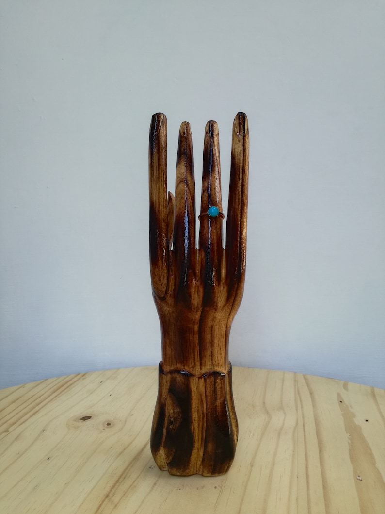 K&ouml;nnte beinhalten: Ein handf&ouml;rmiger Ringhalter aus Holz mit dunkelbraunem Finish. Ein t&uuml;rkisfarbener Ring wird an einem der Finger pr&auml;sentiert. Der Ringhalter steht auf einer hellen Holzoberfl&auml;che.