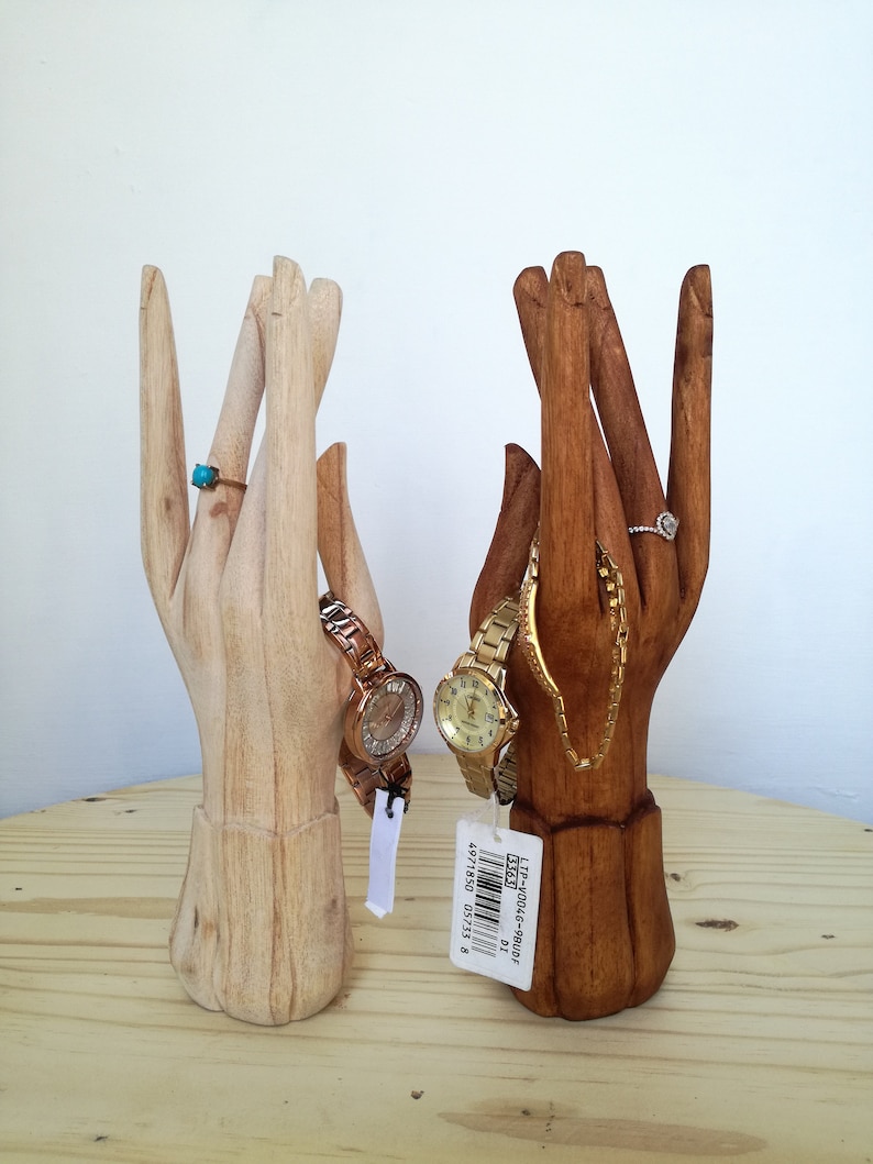 Puede incluir: Dos soportes para joyas de madera con forma de mano. Uno es de madera natural y el otro est&aacute; te&ntilde;ido de marr&oacute;n oscuro. Ambas manos sostienen relojes y anillos.